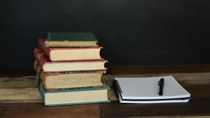 Pile of old hardback books next to a spiral notepad and pen on a rustic wooden table