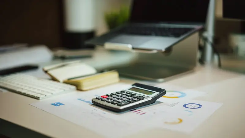 Calculator on printed charts on a desk, with a notebook, keyboard and laptop softly out of focus behind