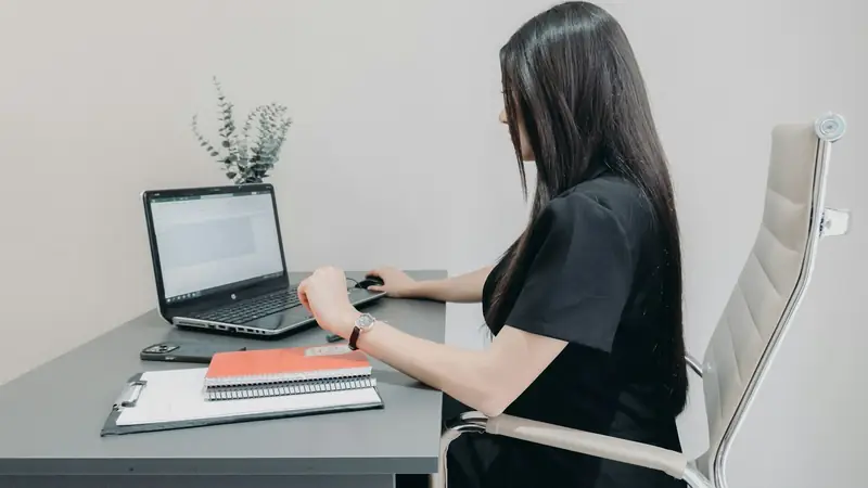 Woman working on a laptop at a grey desk with a clipboard and notebooks beside her in a plain, quiet office