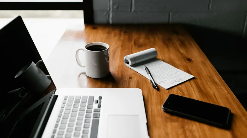 Wooden desk in window light with a laptop, a mug of black coffee, a curled notepad of handwritten notes with a pen, and a phone