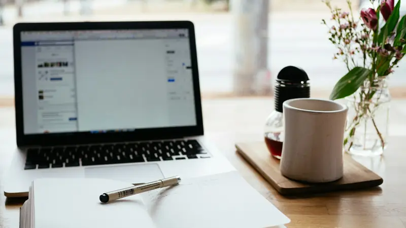 Open paper notebook with a felt-tip pen resting on it, in front of a laptop on a wooden table by a sunny window, with a mug and a jar of pink flowers