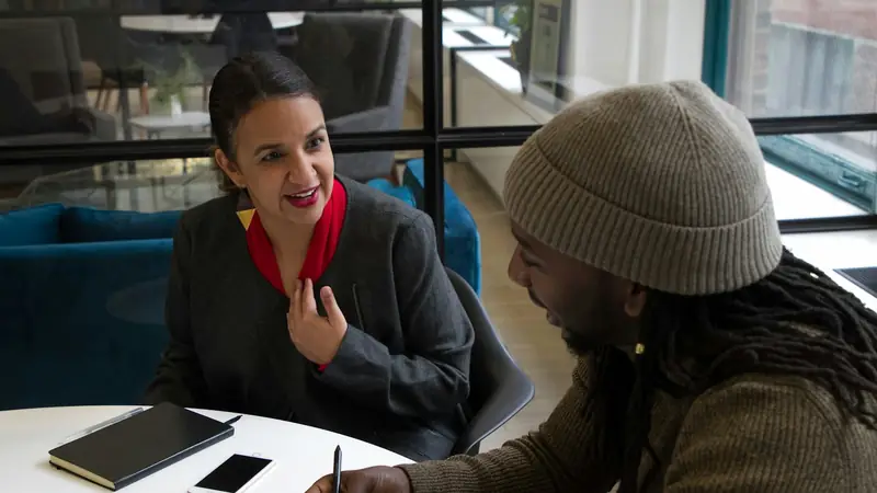 Woman in a red scarf talking with a man in a knitted beanie across a small round table with a notebook and phone