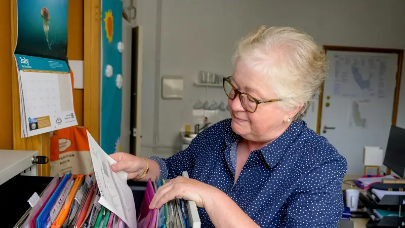 Older woman in glasses leafing through colourful folders in a filing drawer in a small, lived-in office