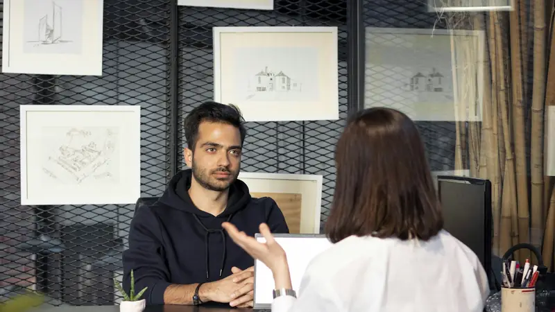 Young man in a hoodie listening across a desk to a woman seen from behind, in an office with framed sketches on the wall