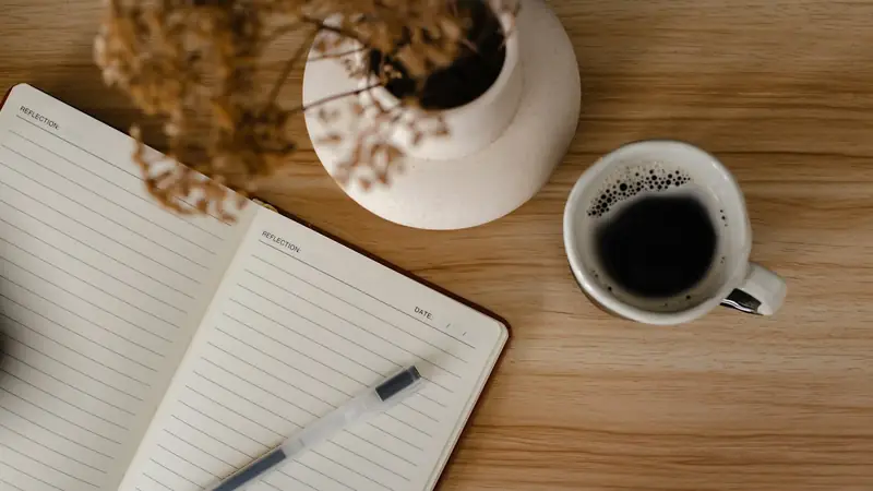 Overhead view of an open lined journal with a pen, a black coffee and a vase of dried flowers on a light oak desk