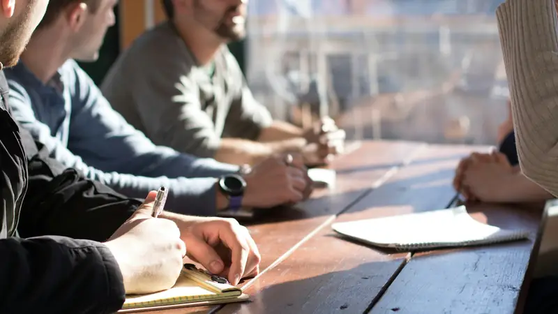 People seated along a sunlit wooden table in a meeting, one in the foreground writing in a notepad