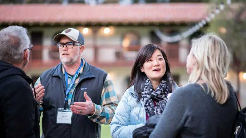 Group of people chatting outdoors at an evening event, a man in a cap and name badge talking with a woman in a scarf