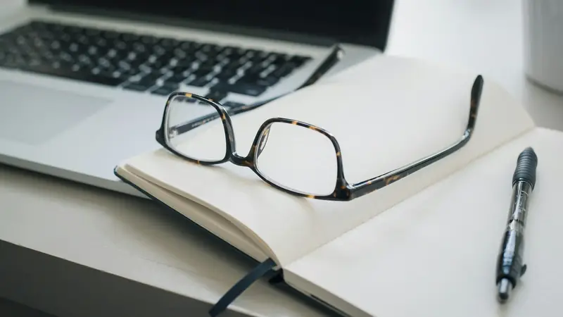 Tortoiseshell reading glasses and a black pen on an open blank notebook, resting on a laptop keyboard