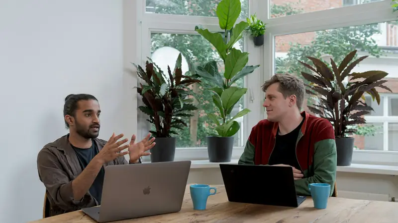 Two men talking across a wooden table by a window full of plants, one explaining with his hands, laptops and blue mugs in front of them
