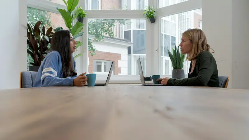Two women sitting opposite each other at the far end of a long wooden table by a large window, talking over laptops and mugs