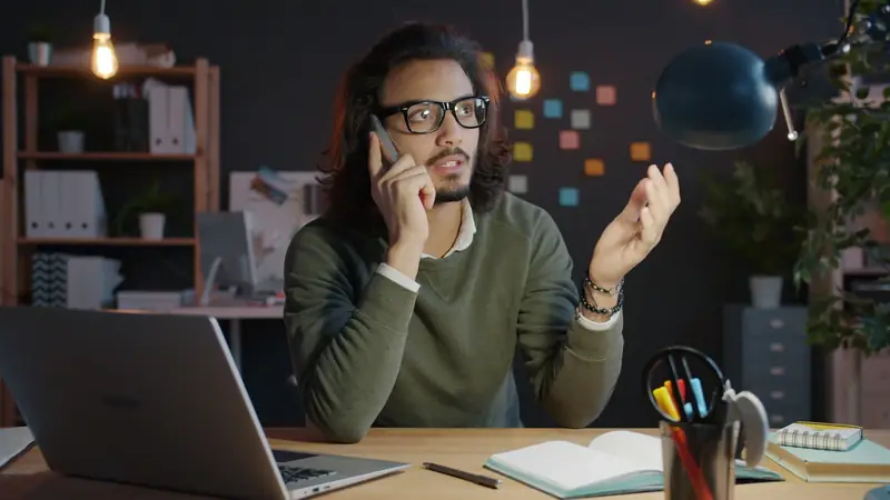 Man in glasses on a phone call at a desk in a warmly lit office, gesturing with his free hand mid-conversation, laptop and notebook in front of him
