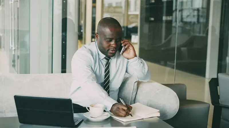 Man in a shirt and tie on a phone call, writing notes in a notebook on a low table beside a coffee and a tablet