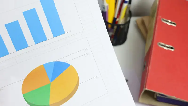 Printed bar and pie charts on paper at an angle beside a red ring binder and a pot of pens