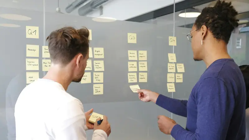 Two colleagues arranging handwritten sticky notes under quarter headings Q1 to Q4 on a glass wall