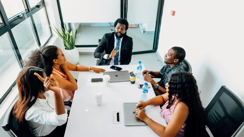 Overhead view of a team of five in a meeting round a white table, one man leading the discussion while others respond